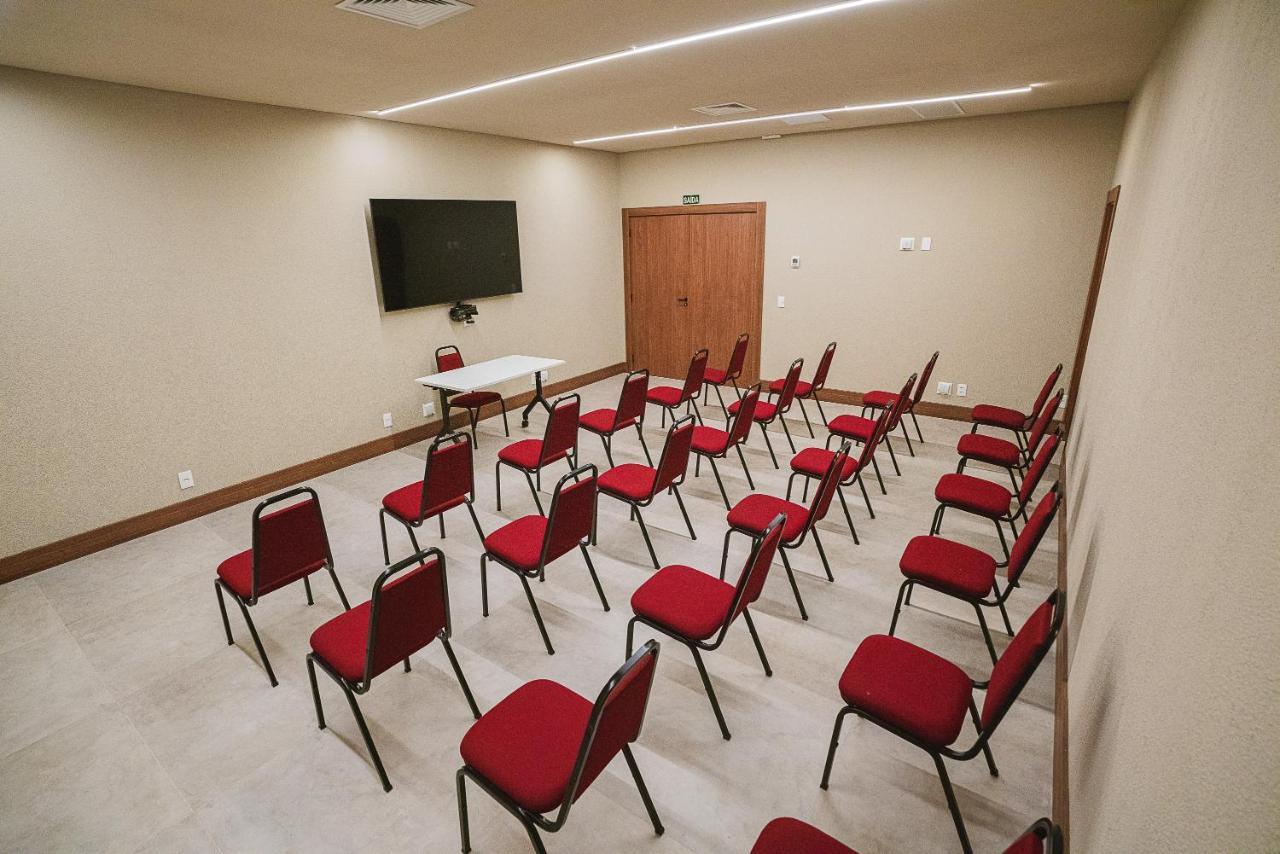 Buona Vitta Gramado Resort & Spa By Gramado Parks エクステリア 写真 The photo shows a conference or meeting room with a simple and modern design. There are several rows of red chairs arranged in a semi-circle facing a white table at the front. On the wall opposite the chairs, there is a flat-screen television mounted