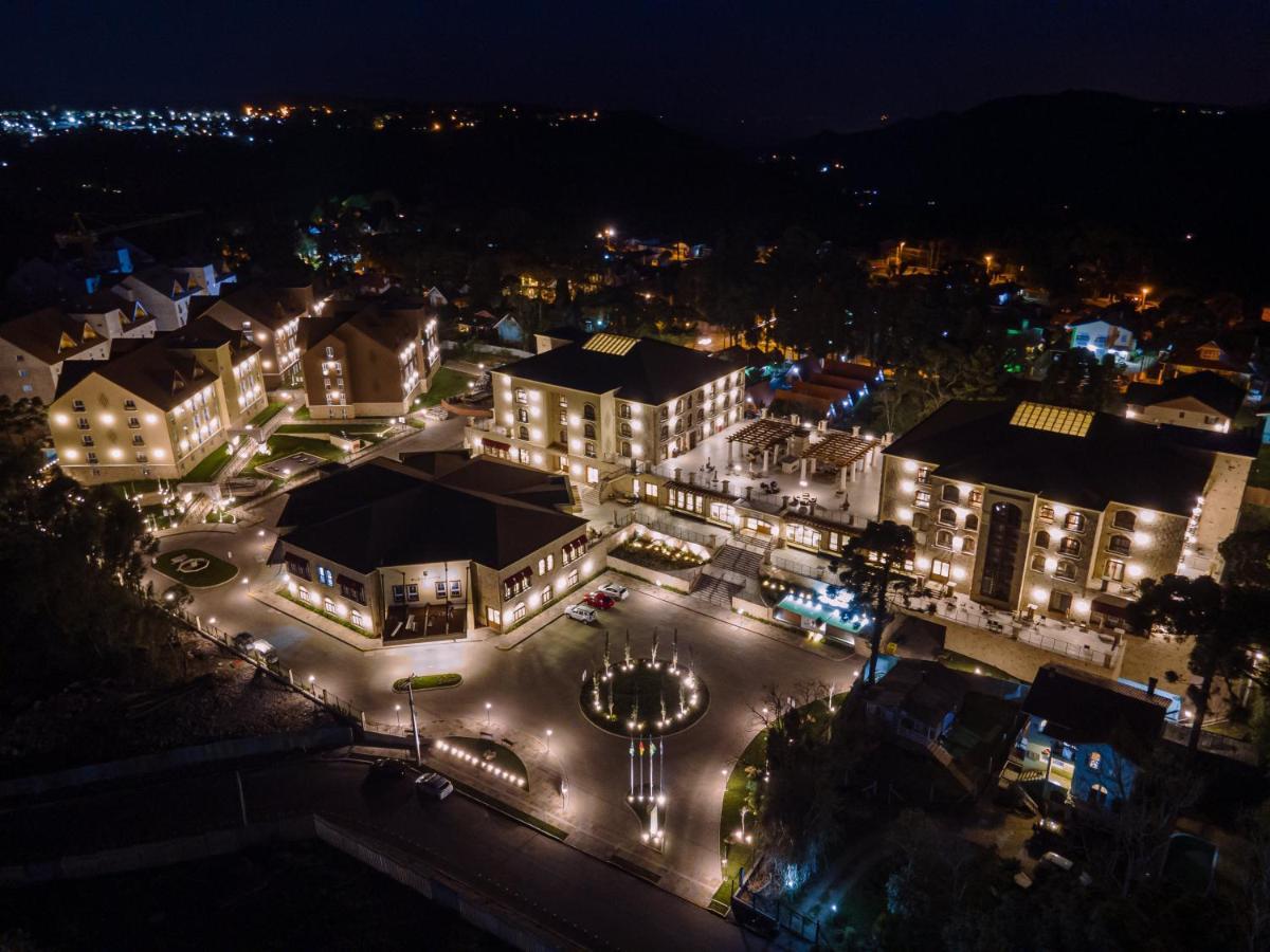 Buona Vitta Gramado Resort & Spa By Gramado Parks エクステリア 写真 The photo shows a beautifully lit resort or hotel complex taken at night. The buildings are well-illuminated, highlighting their architecture. There is a circular area with a fountain at the center, surrounded by pathways that connect various parts o