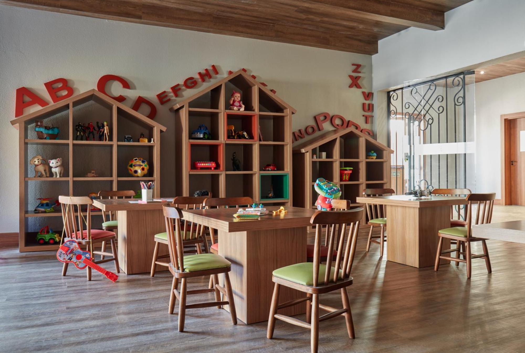 Buona Vitta Gramado Resort & Spa By Gramado Parks エクステリア 写真 The photo shows a classroom or playroom designed for children. It features wooden furniture, including several tables and chairs with green cushions. In the background, there are large, open shelving units shaped like houses, displaying various toys 