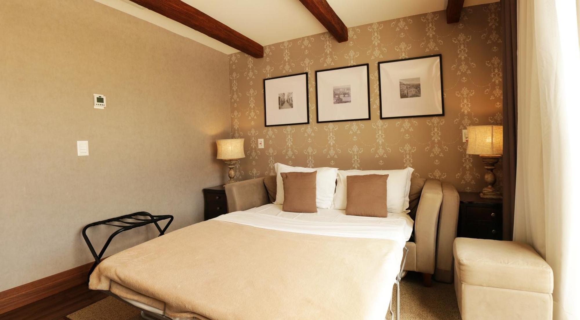 Buona Vitta Gramado Resort & Spa By Gramado Parks エクステリア 写真 The photo shows a cozy bedroom with a neutral color palette. There is a bed with white linens and two brown decorative pillows. On either side of the bed, there are bedside tables with lamps. The walls feature a subtle floral pattern, and three frame