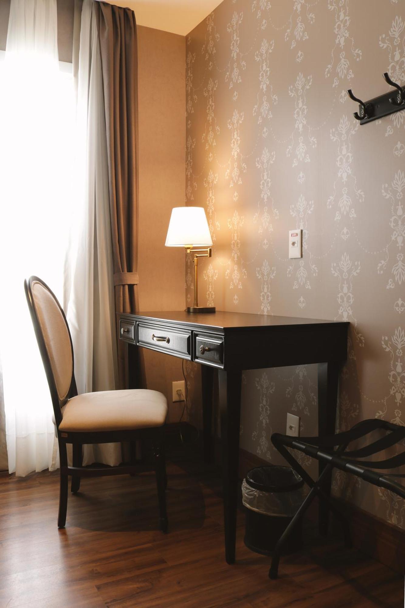 Buona Vitta Gramado Resort & Spa By Gramado Parks エクステリア 写真 The photo shows a small desk set against a wall with a decorative pattern. The desk is black with a few drawers and has a small lamp on it. Next to the desk, there is a chair with a light-colored cushion. Soft light is coming through a window, castin