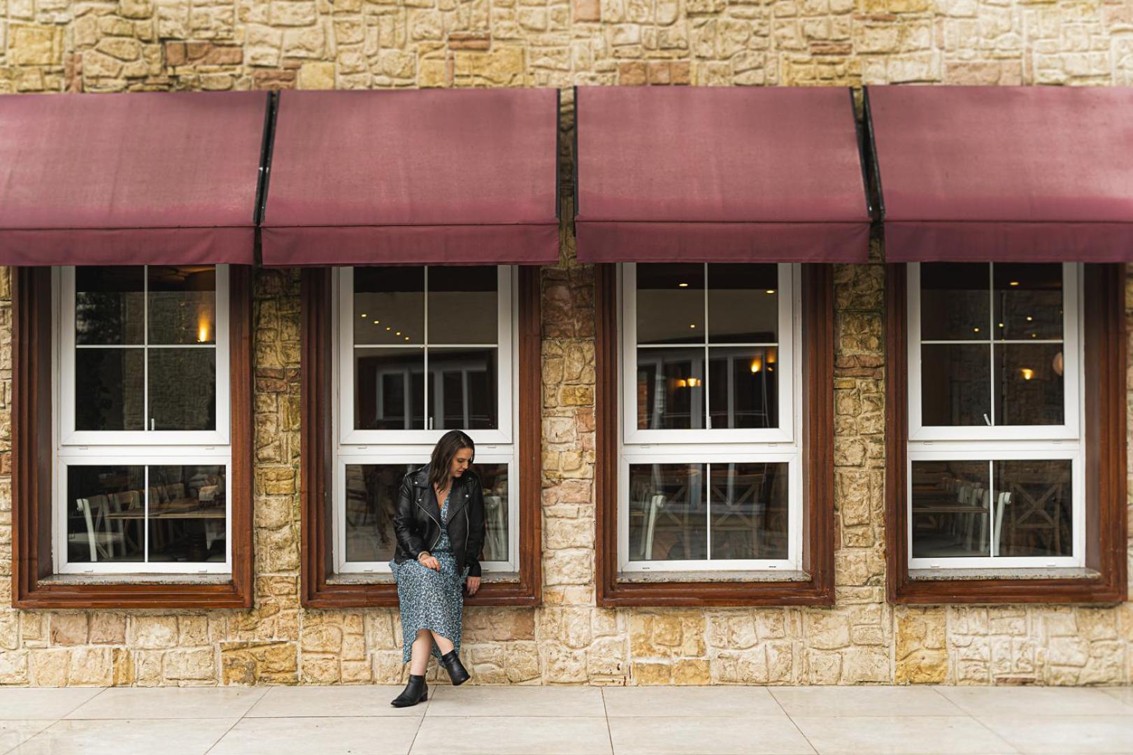 Buona Vitta Gramado Resort & Spa By Gramado Parks エクステリア 写真 The photo shows a woman standing against a stone wall with three large windows in front of her, each framed with wooden borders. The windows have maroon awnings above them. The woman is dressed in a long skirt and leather jacket, leaning slightly aga